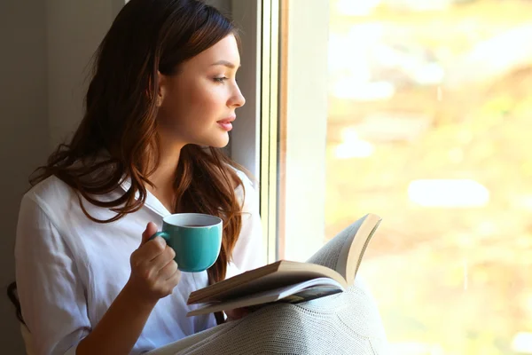 Jonge vrouw thuis zitten in de buurt van raam ontspannen in haar woonkamer lezen boek en het drinken van koffie of thee — Stockfoto