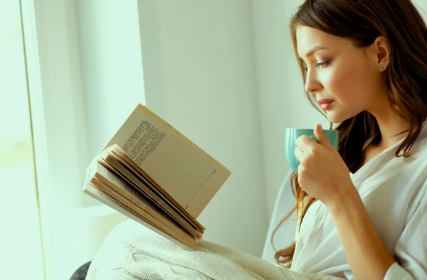 Young woman at home sitting near window relaxing in her living room reading book and drinking coffee or tea