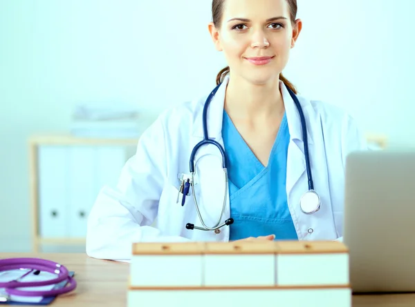 Mooie jonge glimlachende vrouwelijke arts zitten aan het bureau en schrijven. — Stockfoto