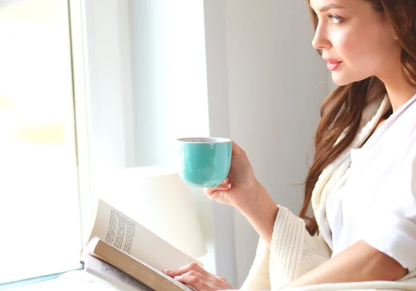 Jonge vrouw thuis zitten in de buurt van raam ontspannen in haar woonkamer lezen boek en het drinken van koffie of thee — Stockfoto