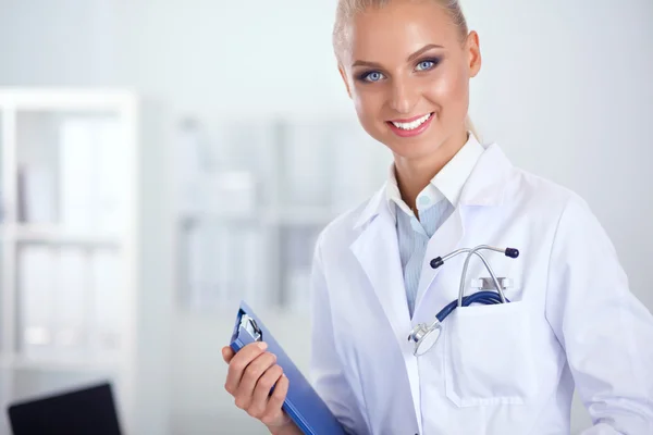 Bonito jovem sorridente médico feminino sentado na mesa e escrevendo. — Fotografia de Stock