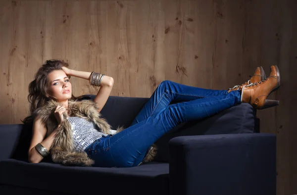 Retrato de mulher elegante sentado no sofá preto vestindo uma calça azul e colete de pele — Fotografia de Stock