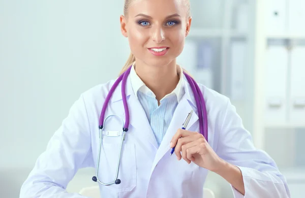 Bonito jovem sorridente médico feminino sentado na mesa e escrevendo. — Fotografia de Stock