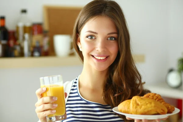 Mujer joven con vaso de jugo y pasteles de pie en la cocina . —  Fotos de Stock