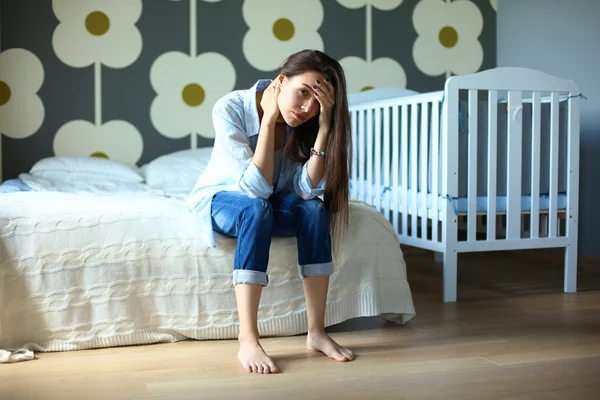 Young tired woman sitting on the bed near childrens cot. — Stock Photo, Image