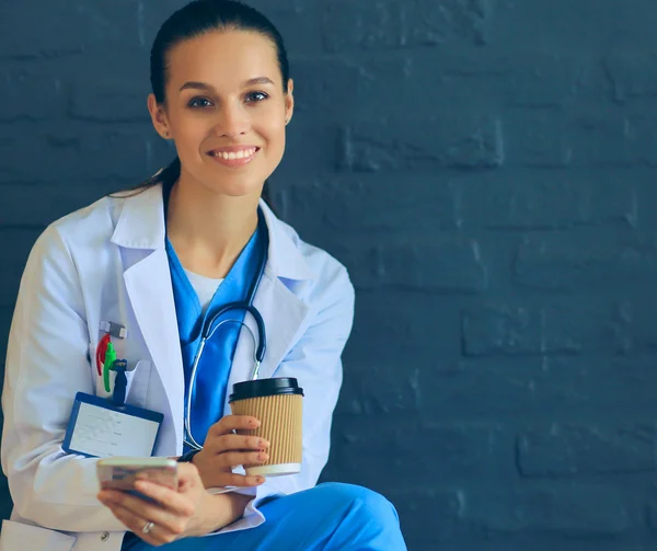 Female doctor sitting with mobile phone and drinking coffee