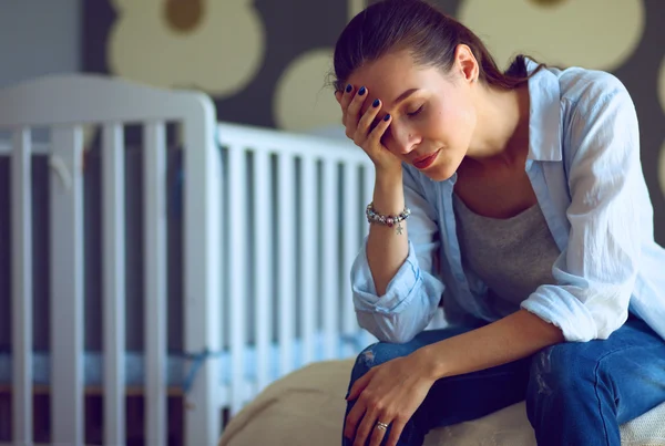 Unga trött kvinna som sitter på sängen nära barnens barnsäng. — Stockfoto