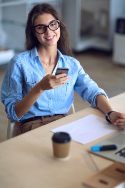 Ofis masasında oturan ve elinde cep telefonu olan genç ve güzel bir iş kadını.