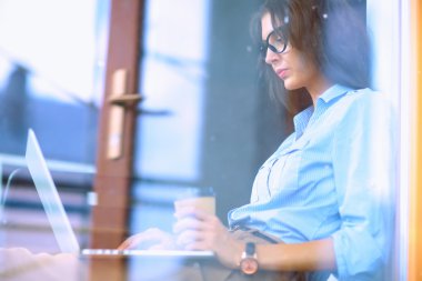 Ofis masasında dizüstü bilgisayarlı genç bir kadın oturuyor, pencereden bakıyor.