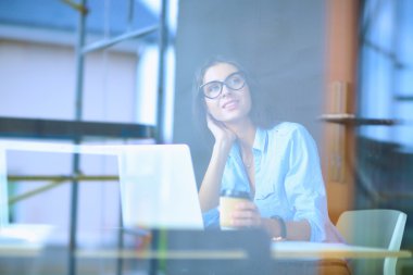 Ofis masasında dizüstü bilgisayarlı genç bir kadın oturuyor, pencereden bakıyor.