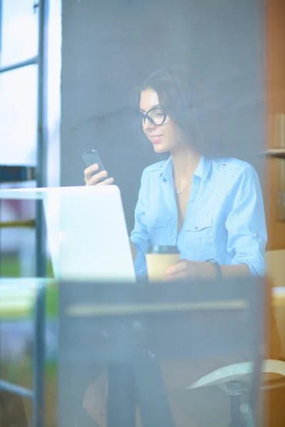 Beautiful young business woman sitting at office desk and holding cell phone, view through window