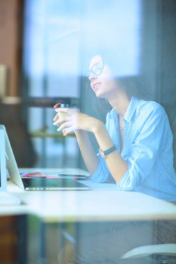 Ofis masasında dizüstü bilgisayarlı genç bir kadın oturuyor, pencereden bakıyor.