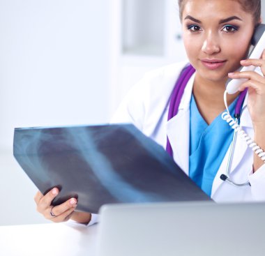 Röntgen taraması yapan kadın doktor teşhis merkezinde telefonla konuşuyor, masada oturuyor.