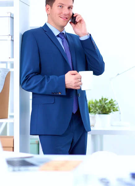 Hombre de negocios sonriente de pie y usando el teléfono móvil en la oficina —  Fotos de Stock