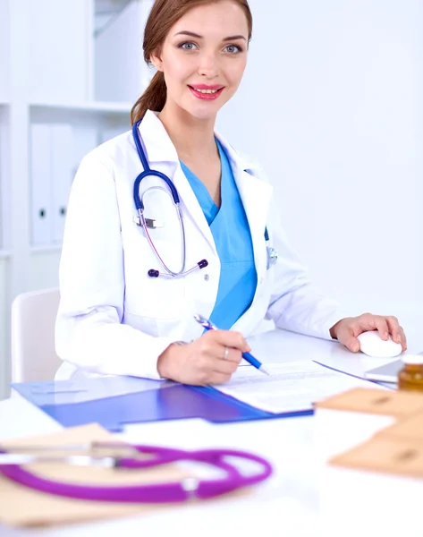 Schöne junge lächelnde Ärztin sitzt am Schreibtisch und schreibt. — Stockfoto