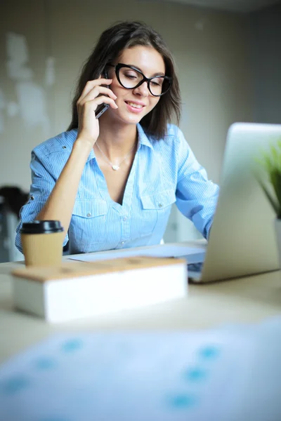 Beautiful young business woman sitting at office desk and talking on cell phone — Stock Photo, Image