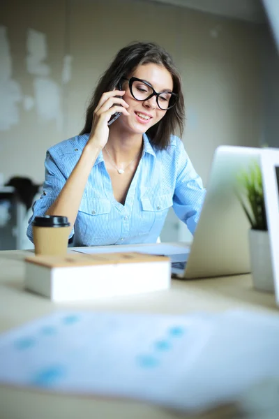 Güzel genç iş kadını ofis masasında oturuyor ve cep telefonuyla konuşuyor. — Stok fotoğraf