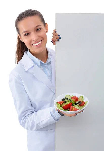 Portrait of a beautiful woman doctor holding a plate with fresh vegetables standing near blank — Stock Photo, Image