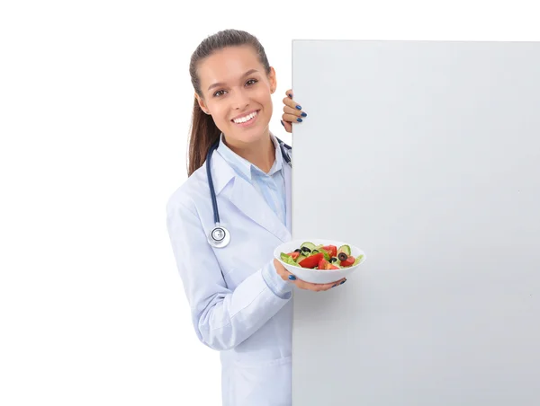 Portrait of a beautiful woman doctor holding a plate with fresh vegetables standing near blank — Stock Photo, Image