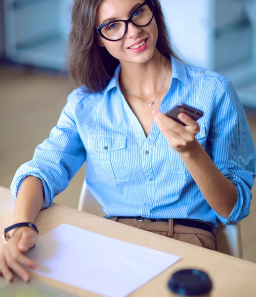 Mulher de negócios jovem bonita sentada na mesa do escritório e segurando telefone celular — Fotografia de Stock