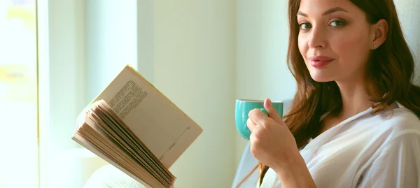 Junge Frau sitzt zu Hause am Fenster und entspannt in ihrem Wohnzimmer, liest Buch und trinkt Kaffee oder Tee — Stockfoto