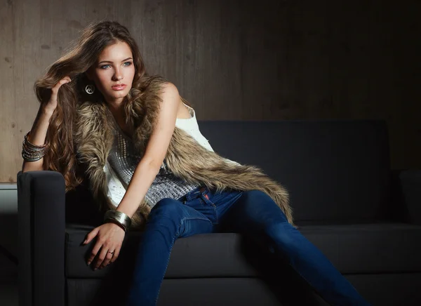 Portrait of elegant woman sitting on black sofa wearing a blue jeans and fur vest — Stock Photo, Image