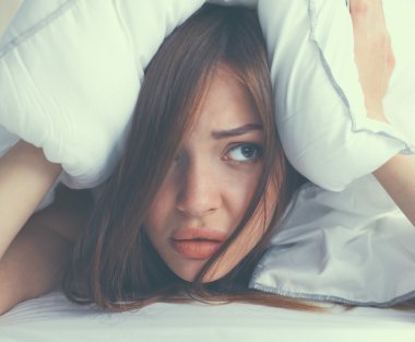 Kadın yatakta uzanıyor ve kulaklarını yastıkla kapatıyor.