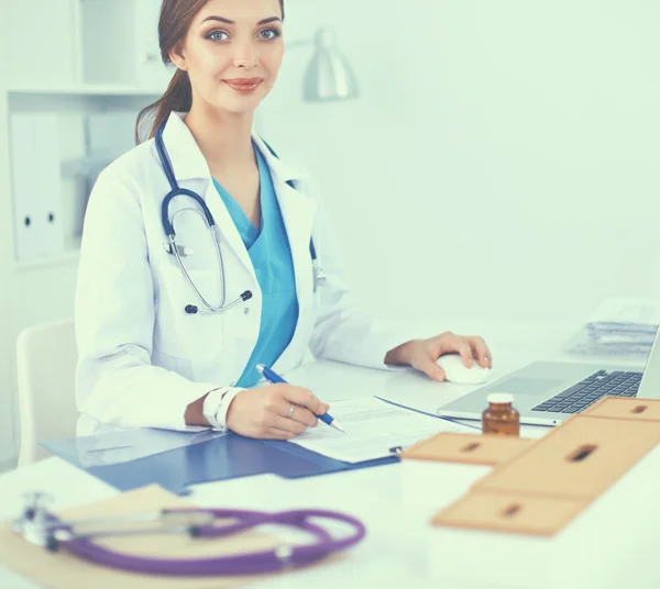 Schöne junge lächelnde Ärztin sitzt am Schreibtisch und schreibt. — Stockfoto