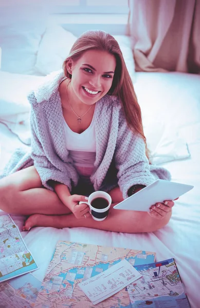 Avslappnad ung kvinna sitter på sängen med en kopp kaffe och digital tablett — Stockfoto