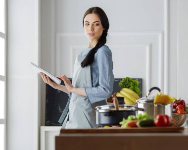 Mutfakta yemek yapmak için tablet bilgisayar kullanan genç bir kadın.