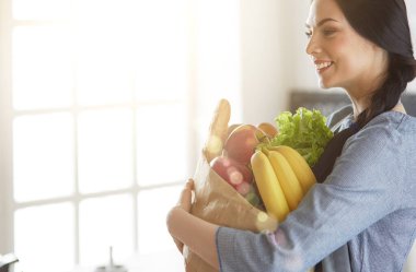 Sebzeli alışveriş poşeti taşıyan genç bir kadın. Mutfakta duruyor.