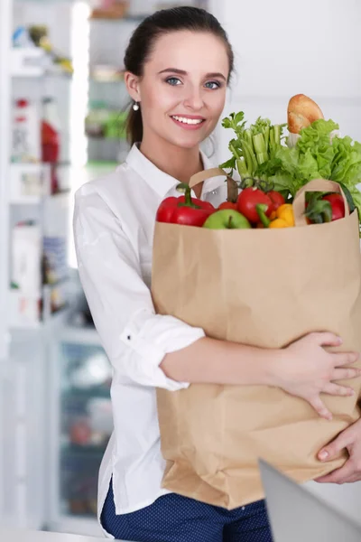 野菜と食料品の買い物袋を持っている若い女性.キッチンに立って.台所の女性がカメラを見て — ストック写真