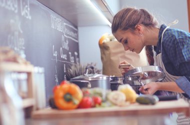 Genç bir kadın mutfağında yemek pişiriyor sobanın yanında duruyor.