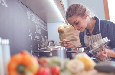 Genç bir kadın mutfağında yemek pişiriyor sobanın yanında duruyor.