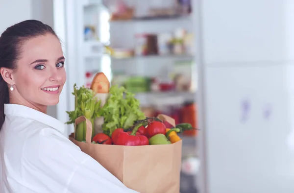 Mujer joven sosteniendo bolsa de la compra de comestibles con verduras.De pie en la cocina. Mujer en la cocina mirando a la cámara — Foto de Stock