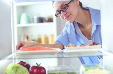 Sağlıklı yiyecek, sebze ve meyvelerle dolu açık buzdolabının yanında duran kadın portresi..