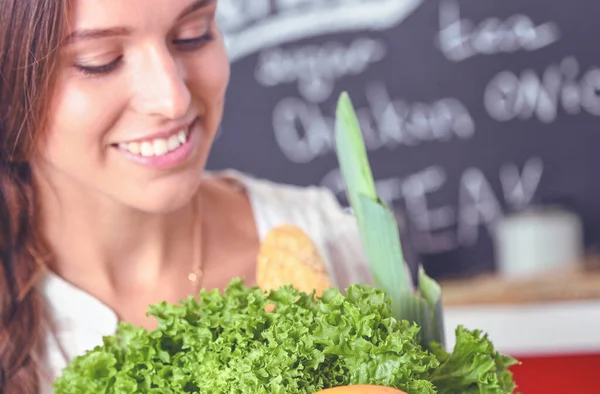 Mujer joven sonriente sosteniendo verduras de pie en la cocina. Jovencita sonriente —  Fotos de Stock