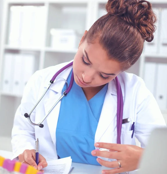 Mooie jonge glimlachende vrouwelijke arts zitten aan het bureau en schrijven. — Stockfoto