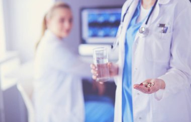 Doktorun arka planda meslektaşıyla klinikte elinde haplar ve bir bardak su tutarken görüntüsü..