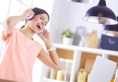 Mutfakta dururken kulaklıkla müzik dinleyen ve dizüstü bilgisayar kullanan neşeli bir genç kadının portresi