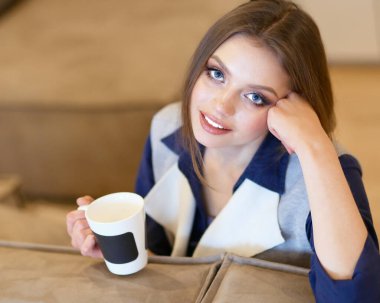 Genç bir kadın, beyaz odada rahat bir koltukta, elinde kahveyle oturuyor.