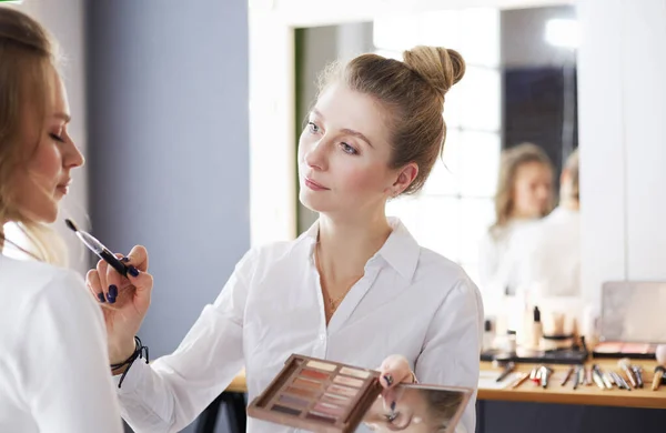 Make-up artist applicando ombretto bianco nell'angolo dei modelli occhio e tenendo una conchiglia con ombretto sullo sfondo — Foto Stock