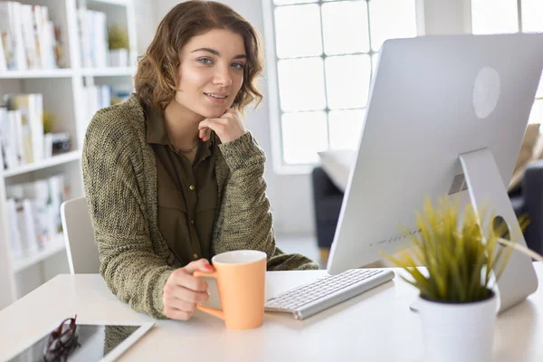 Mulher sorridente atraente sentado na mesa do escritório, segurando uma xícara de café, ela está relaxando e olhando para longe — Fotografia de Stock