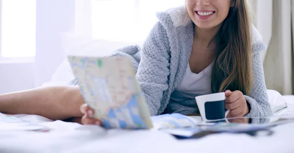 Avslappnad ung kvinna sitter på sängen med en kopp kaffe och digital tablett — Stockfoto