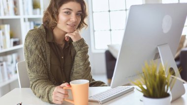 Çekici, gülümseyen bir kadın büro masasında oturuyor, elinde bir fincan kahve tutuyor, rahatlıyor ve başka tarafa bakıyor.