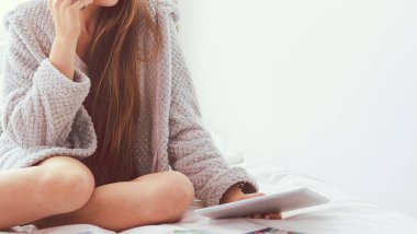 Bir fincan kahve ve dijital tabletle yatakta oturan rahat genç bir kadın.