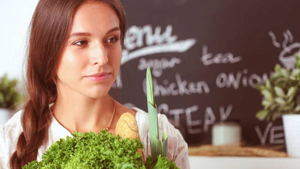 Mujer joven sonriente sosteniendo verduras de pie en la cocina. Jovencita sonriente —  Fotos de Stock