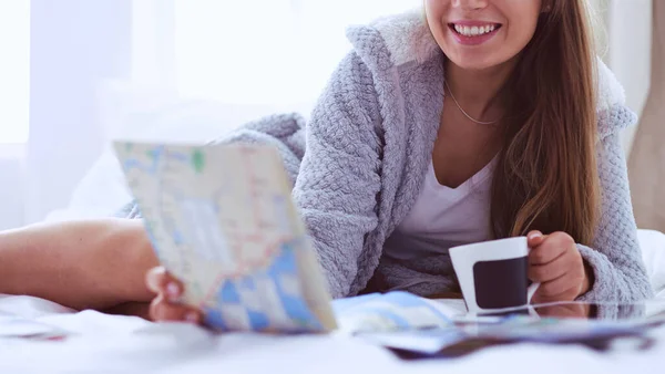 Avslappnad ung kvinna sitter på sängen med en kopp kaffe och digital tablett — Stockfoto