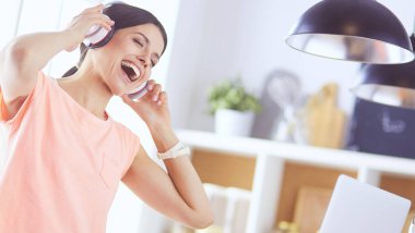 Mutfakta dururken kulaklıkla müzik dinleyen ve dizüstü bilgisayar kullanan neşeli bir genç kadının portresi