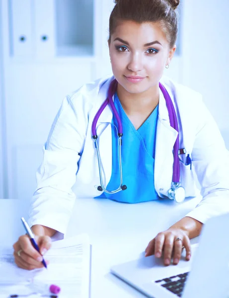 Hermosa joven sonriente doctora sentada en el escritorio y escribiendo. —  Fotos de Stock